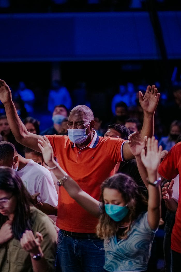 A diverse group of masked individuals raising hands in prayer during a worship service.