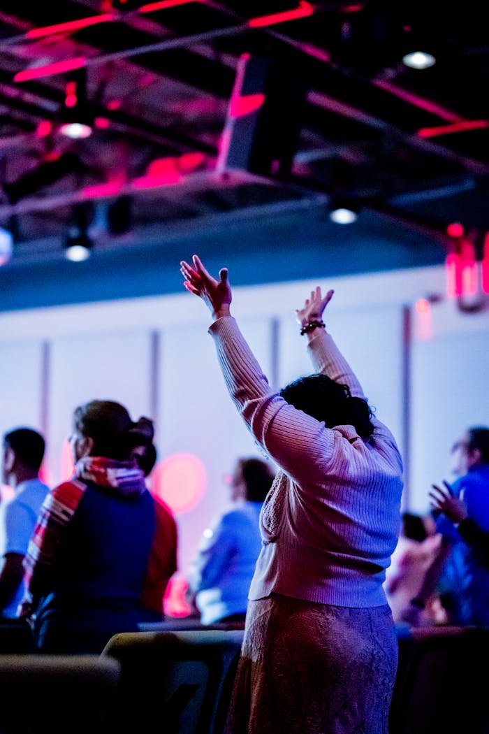 who-we-are A group of diverse people raising hands in worship in a vibrant indoor setting under colorful lighting.